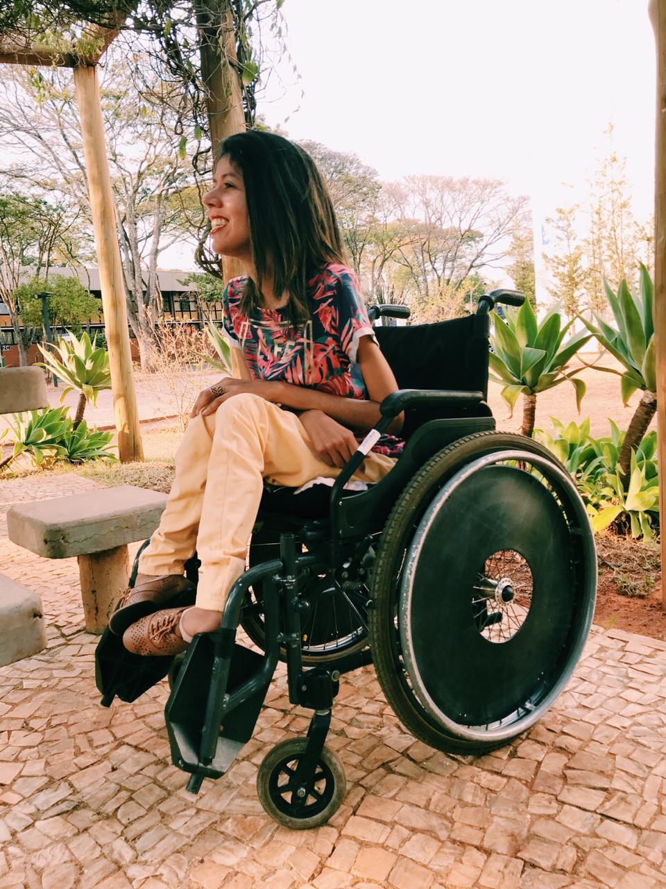 Foto da Fatine Oliveira, ela tem cabelos castanhos até o ombro, veste uma blusa estampada, uma calça amarela e sapatos marrons, sentada em uma cadeira de rodas. Ao fundo, um jardim.