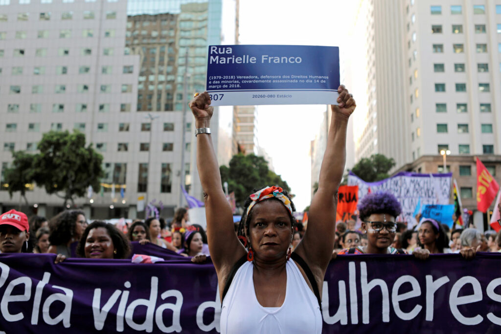 mulher negra segura placa escrito rua marielle franco, ao fundo um grupo grande de mulheres segura uma faixa escrito "pela vida das mulheres"